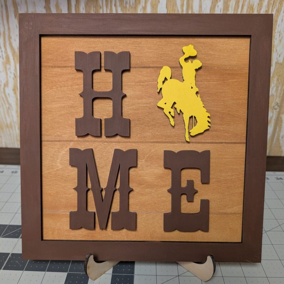Red Rooster Laser Arts The DIY Small Wooden Steamboat Home Sign features large brown letters spelling HOME, with the O replaced by a yellow cowboy silhouette. Framed and perfect for display on a small easel, it complements any DIY wooden bee-themed welcome sign kit.
