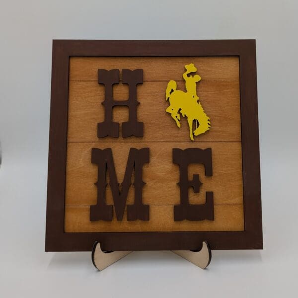 Red Rooster Laser Arts The DIY Wooden Wyoming Tic Tac Toe Kit features dark brown letters spelling HOME, with a yellow cowboy silhouette as the O. The set is displayed on a small easel against a plain background.