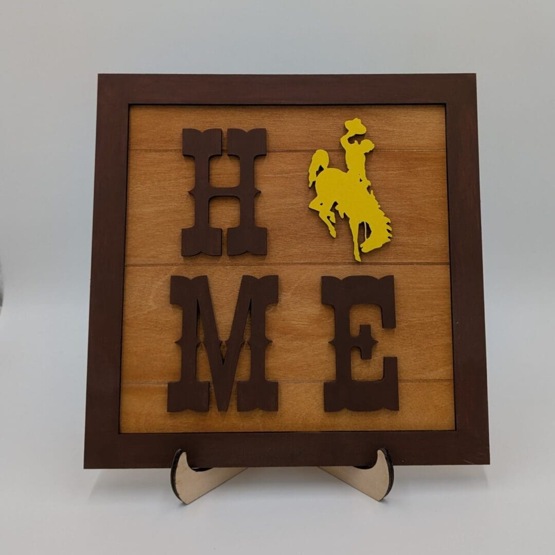 Red Rooster Laser Arts The DIY Wooden Wyoming Tic Tac Toe Kit features dark brown letters spelling HOME, with a yellow cowboy silhouette as the O. The set is displayed on a small easel against a plain background.