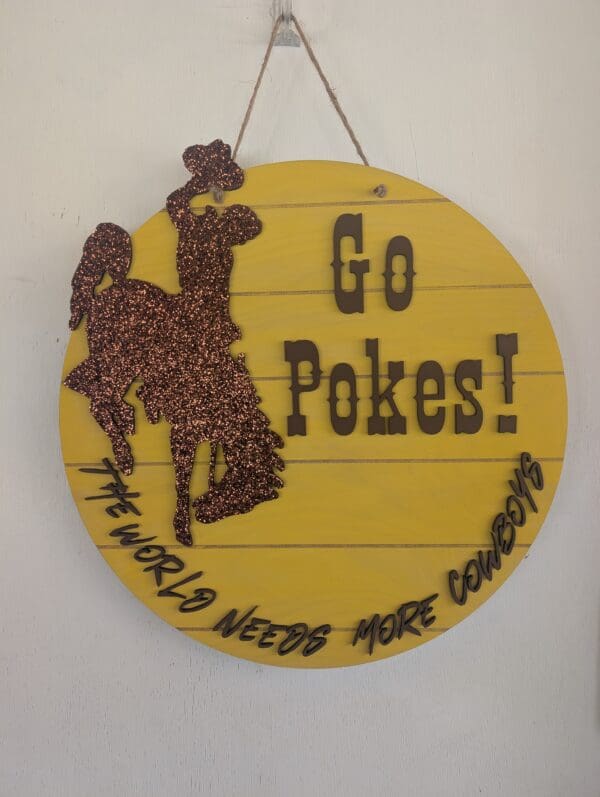 Red Rooster Laser Arts The DIY Door Hanger Kit - University of Wyoming Pride includes a yellow wooden sign with a glittery brown cowboy on a bucking horse and bold black text: Go Pokes! and The world needs more cowboys. Perfect for crafting fans!.