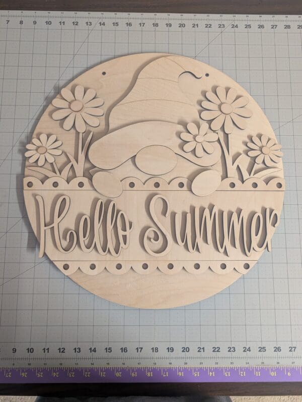 Red Rooster Laser Arts The DIY Door Hanger Kit - Gnome Summer features a round wooden sign with Hello Summer in cursive and a gnome in a hat surrounded by flowers, displayed on a gridded cutting mat with rulers along the edges.