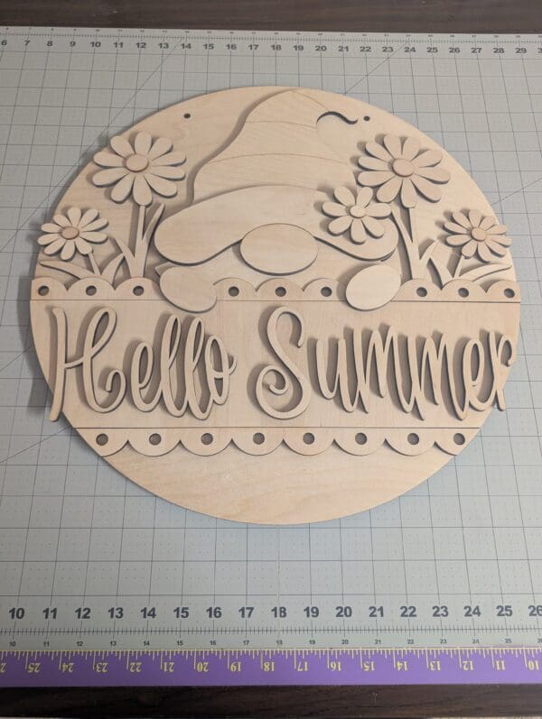 Red Rooster Laser Arts The DIY Door Hanger Kit - Gnome Summer includes a round, unfinished wooden sign with a gnome, flowers, and Hello Summer in decorative script—ideal for creating your own summer door hanger. Shown on a cutting mat with measuring gridlines.