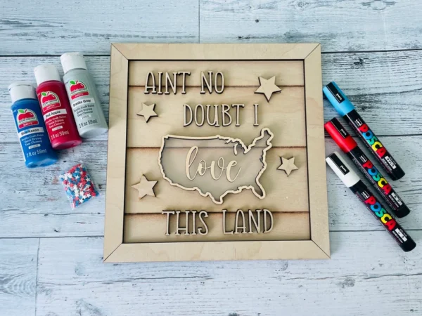 Red Rooster Laser Arts The DIY Patriotic Shaker Sign Kit - Love This Land features a U.S. map cutout with “AINT NO DOUBT I love THIS LAND,” star accents, paint bottles, markers, and star confetti on light wood.
