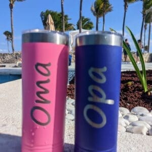 Red Rooster Laser Arts Two insulated tumblers rest poolside. The pink one reads Oma, and the blue one reads Opa, highlighting the charm of personalized products. Palm trees sway in the background under a clear blue sky. This can be personalized for you by Red Rooster Laser Arts.