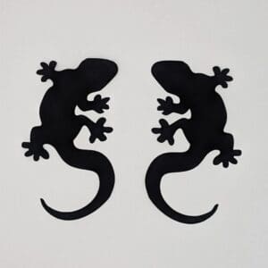 Red Rooster Laser Arts Two pre-fused, laser-cut black gecko silhouettes from the Set of 2 Geckos face each other on a white background. Their legs are spread with five splayed toes per foot, and upward-curling tails add to their harmony. These are laser cut and pre fused appliques, made from high quality quilting cotton.