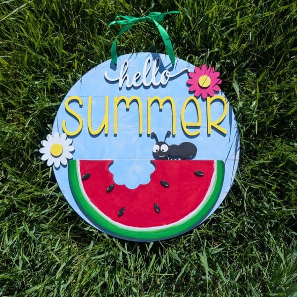 Red Rooster Laser Arts The DIY Door Hanger Kit - Welcome Summer features a round sign on grass with hello summer in yellow, painted flowers, an ant, and a watermelon slice. It includes a green hanging ribbon and blue sky background—perfect as a summer decor project.