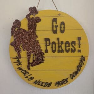 Red Rooster Laser Arts The DIY Door Hanger Kit - University of Wyoming Pride includes a yellow wooden sign with a glittery brown cowboy on a bucking horse and bold black text: Go Pokes! and The world needs more cowboys. Perfect for crafting fans!.
