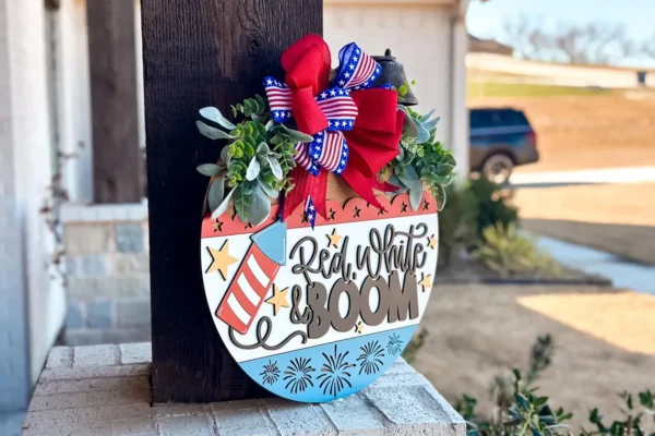 Red Rooster Laser Arts A badge-shaped DIY Door Hanger Kit - Red, White and BOOM! features greenery, a red, white, and blue bow, and Red, White & Boom—the perfect patriotic decor for your porch.