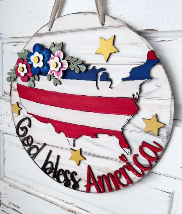 Red Rooster Laser Arts The DIY Door Hanger Kit - God Bless America features a round wooden hanger with a rustic white background, a U.S. map in red, white, and blue stripes, gold stars, floral accents, and God bless America in bold letters.