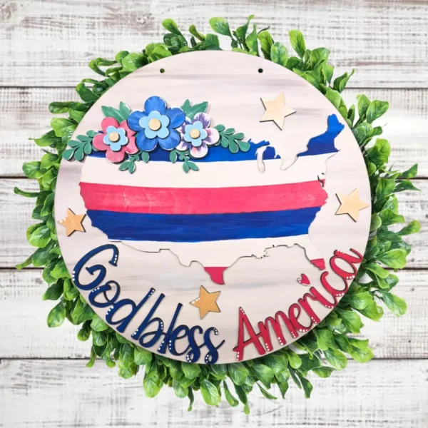 Red Rooster Laser Arts The DIY Door Hanger Kit - God Bless America features a round wreath with green leaves, a wooden U.S. map in red, white, and blue stripes, blue flowers and stars, and “God bless America”—perfect for patriotic décor.