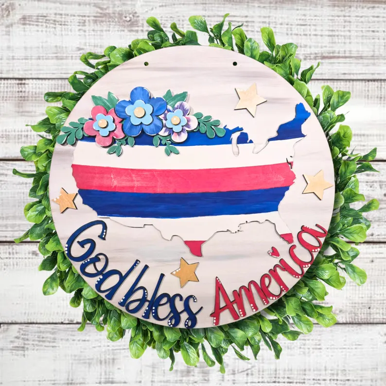 Red Rooster Laser Arts The DIY Door Hanger Kit - God Bless America features a round wreath with green leaves, a wooden U.S. map in red, white, and blue stripes, blue flowers and stars, and “God bless America”—perfect for patriotic décor.