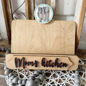 Red Rooster Laser Arts A wooden cookbook holder with an interchangeable Moms Kitchen insert and round utensil emblem sits on a wooden surface, decorated with gray and white beaded garland and a festive DIY CBH Happy Fall Yall insert kit.