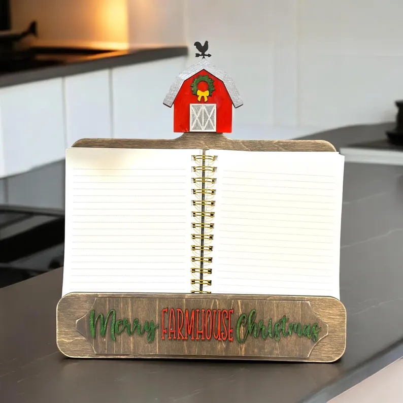 Red Rooster Laser Arts An open spiral notebook sits on a wooden stand with the “Farmhouse Christmas” Interchangeable Cookbook Holder Insert, featuring green and red text and a small red barn—perfect for displaying your CBH Baking Spirits Bright kit on the kitchen counter.