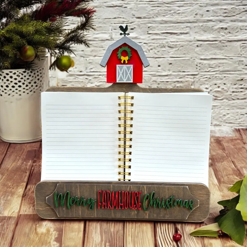 Red Rooster Laser Arts A spiral-bound Interchangeable Cookbook Holder Insert - Farmhouse Christmas rests on a wooden stand with Merry Farmhouse Christmas and a red barn decor, accented by pine branches and apples on a rustic background.