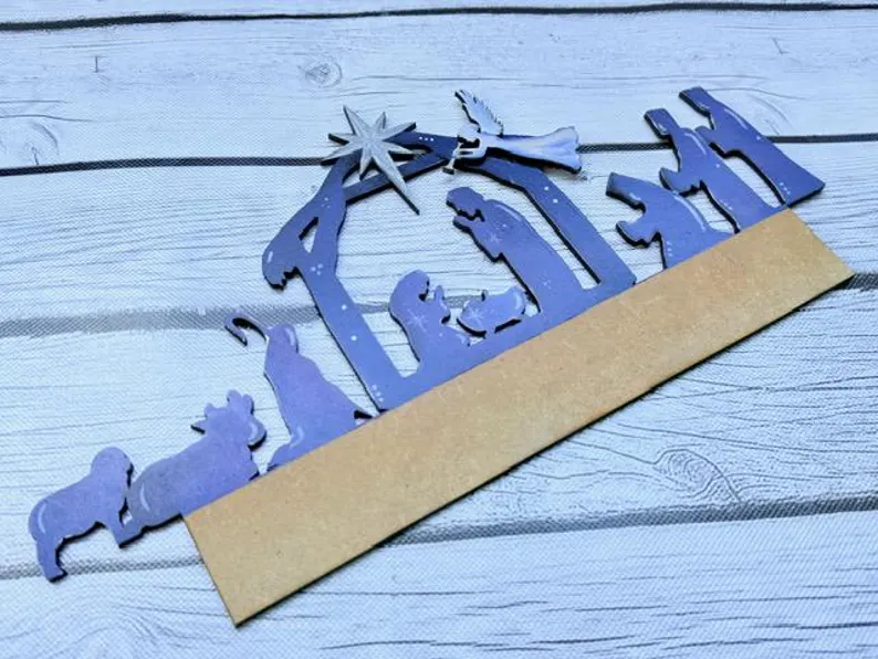 Red Rooster Laser Arts The Interchangeable Door Hanger Insert - Nativity Scene is a purple paper cutout featuring the Holy Family, angel, animals, and Star of Bethlehem on brown cardboard atop a wooden surface—perfect for DIY Christmas décor.
