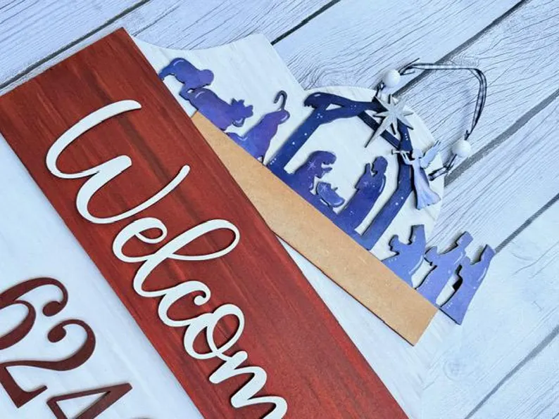 Red Rooster Laser Arts The Interchangeable Door Hanger Insert - Nativity Scene is a wooden welcome sign with a red and white background, featuring a blue nativity silhouette of Mary, Joseph, baby Jesus, wise men, and animals—ideal for DIY Christmas decor.
