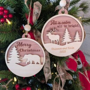 Red Rooster Laser Arts Two wooden ornaments from the Personalized Woodland Ornament Set hang on a tree. One says “Merry Christmas” with a bear; the other reads “All is calm, all is bright,” with a moose and “Shannon and Kevin 2025,” both with festive ribbons.