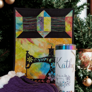 Red Rooster Laser Arts A colorful quilted wall hanging, a tumbler with “Kathy” and a sewing machine design, and a pair of purple knit socks from our Gift Bundle are displayed in front of a decorated Christmas tree with gold ornaments.