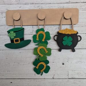 Red Rooster Laser Arts Three St. Patrick’s Day decorations—a leprechaun hat, gold horseshoes with shamrocks, and a pot of gold—hang on the DIY Interchangeable Wall Peg System Decor - St. Patrick's Day, set against a white wood wall for festive DIY style.