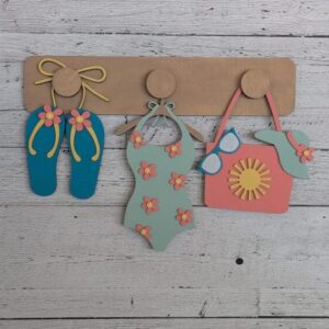 Red Rooster Laser Arts The DIY Interchangeable Wall Peg System Decor - Summer features a light wood hook displaying blue flip-flop, floral swimsuit, and pink beach bag cutouts with spring flowers—ideal for adding a fresh, customizable touch to your space.