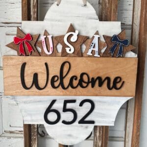 Red Rooster Laser Arts The Interchangeable Door Hanger Insert - USA Stars and Bows features a sign reading USA, Welcome, and 652 on wooden stars with red, white, and blue bows—ideal as a rustic DIY Christmas Tacky Sweater door hanger insert kit.