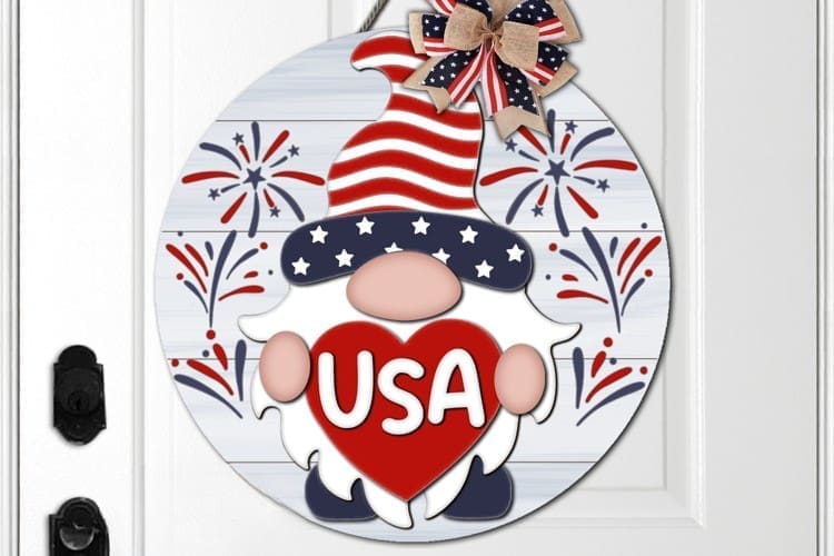 Red Rooster Laser Arts The DIY Door Hanger Kit - "Patriotic USA Gnome" features a round sign with a gnome in a stars-and-stripes hat holding a red heart labeled USA, fireworks background, and an American flag bow at the top.