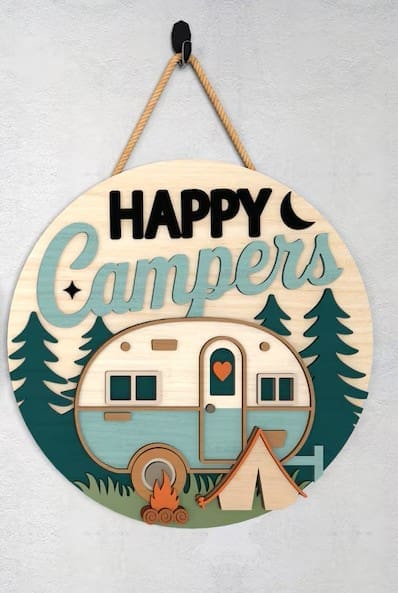 Red Rooster Laser Arts The DIY Door Hanger Kit - "Happy Campers 1" is a round wooden sign with a camper van, tent, pine trees, campfire, and crescent moon design. It features "Happy Campers" text and hangs by rope on a light wall.