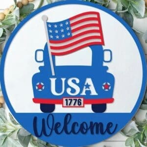 Red Rooster Laser Arts The DIY Door Hanger Kit - "Patriotic Welcome - Truck" features a round sign with a blue truck carrying an American flag, USA on the truck, 1776 on the license plate, “Welcome” below, and green leaves accenting this festive patriotic decor.