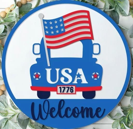Red Rooster Laser Arts The DIY Door Hanger Kit - "Patriotic Welcome - Truck" features a round sign with a blue truck carrying an American flag, USA on the truck, 1776 on the license plate, “Welcome” below, and green leaves accenting this festive patriotic decor.