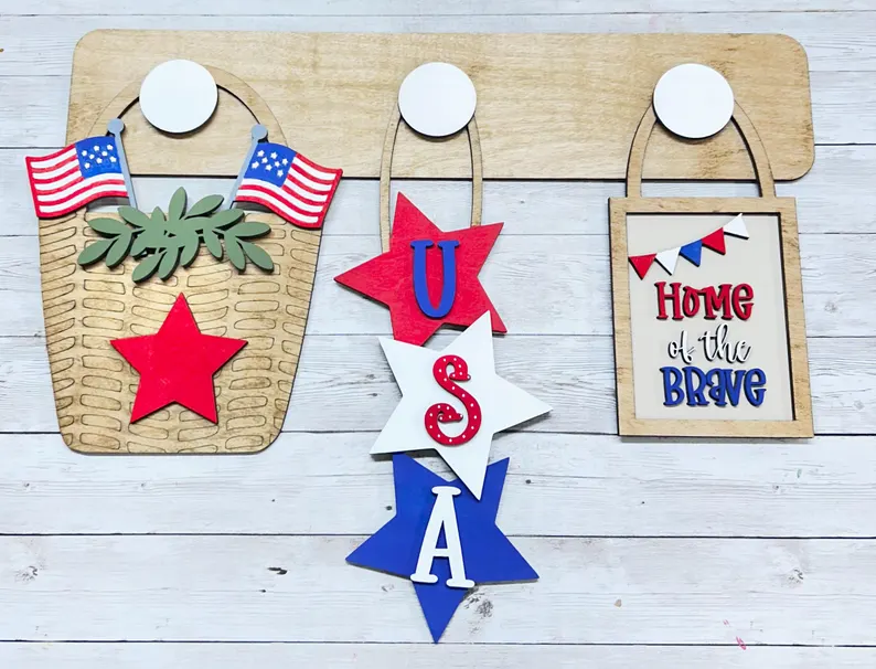 Red Rooster Laser Arts The DIY Interchangeable Wall Peg System Decor - Patriotic Collection includes a wooden base with a basket of American flags, three stars spelling USA, and a "Home of the Brave" sign in festive red, white, and blue accents.
