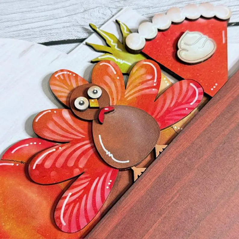 Red Rooster Laser Arts A colorful cartoon turkey and a slice of pumpkin pie with whipped cream are illustrated on paper, partially tucked behind a wooden surface—perfect for accenting your Interchangeable Door Hanger Insert - Fall Coffee Cups.