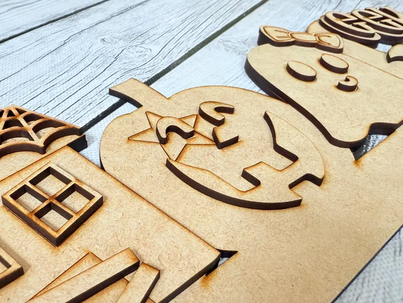 Red Rooster Laser Arts Laser-cut wooden Halloween decorations—a house, a jack-o-lantern with a bow, and a ghost with intricate carved details—arranged on a light wooden surface, make the perfect accents or companions for your Interchangeable Door Hanger Insert - Thanksgiving Harvest (Copy) kit.