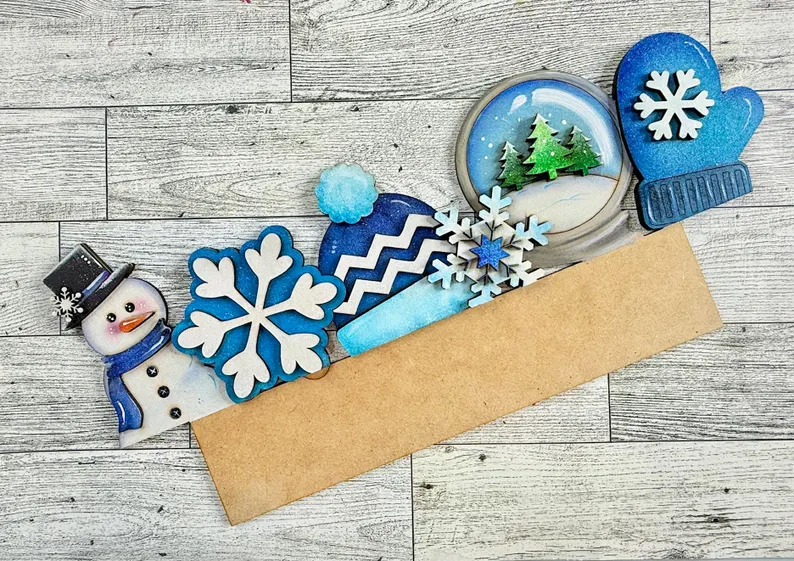 Red Rooster Laser Arts Winter-themed decorations—including a snowman, snowflakes, a cozy hat, a winter scene in a dome, and a mitten—are all arranged above a blank brown rectangular sign on a wooden surface. Perfect for adding to your Interchangeable Door Hanger Insert - Winter Wonderland kit from Red Rooster Laser Arts.