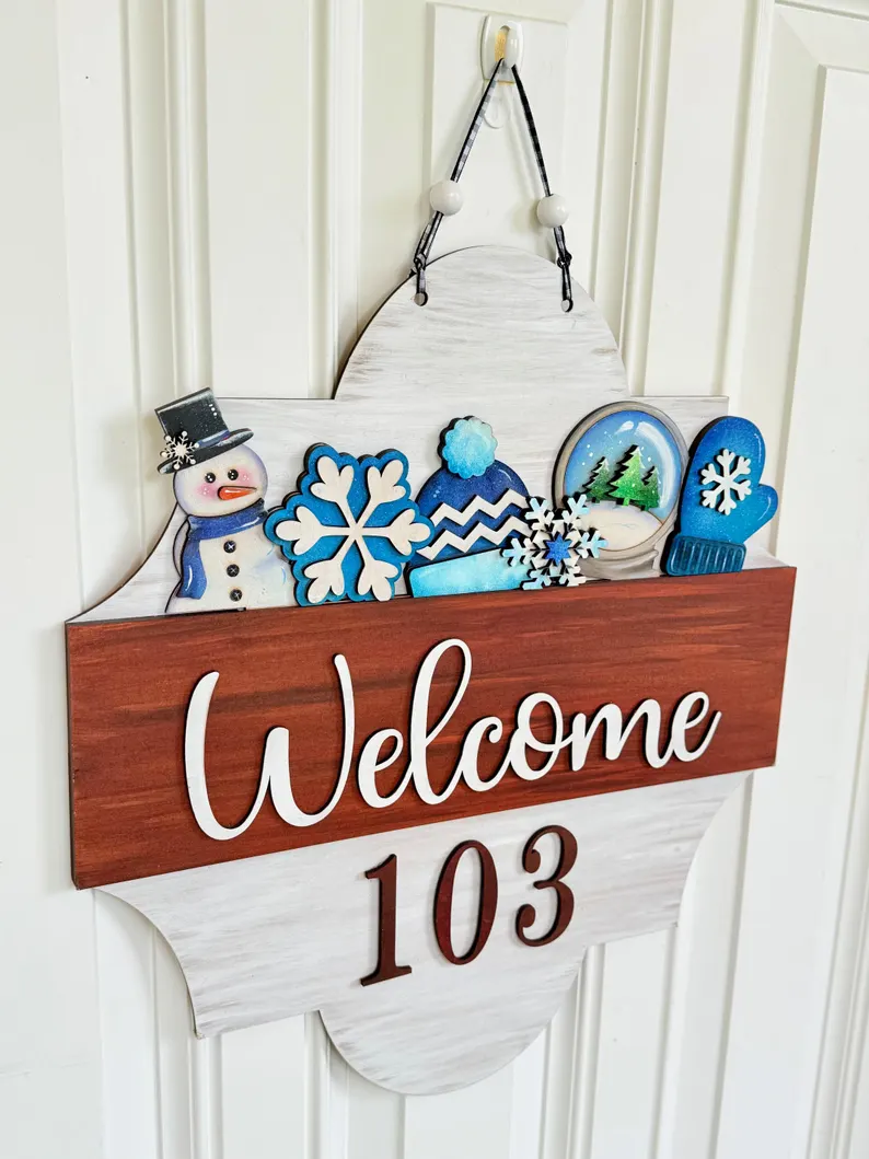 Red Rooster Laser Arts A door sign reads Welcome 103 with winter-themed decorations, including a snowman, snowflakes, a hat, mittens, and a snowy landscape, hanging on a white paneled door. This charming display can be easily created using the Interchangeable Door Hanger Insert - Winter Wonderland from Red Rooster Laser Arts.