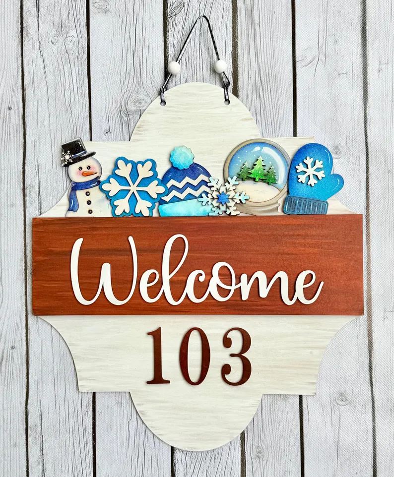 Red Rooster Laser Arts A wooden welcome sign with Welcome 103 and winter decorations—featuring a snowman, snowflakes, mitten, hat, and a snowy scene—hangs on a rustic wood background, making it the perfect match for the Interchangeable Door Hanger Insert - Winter Wonderland from Red Rooster Laser Arts.