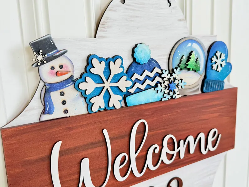 Red Rooster Laser Arts Close-up of a winter-themed wooden welcome sign featuring a snowman, snowflakes, a hat, a mitten, and a snowy landscape—all in blue and white accents above the word "Welcome." This charming décor piece is the Interchangeable Door Hanger Insert - Winter Wonderland from Red Rooster Laser Arts.
