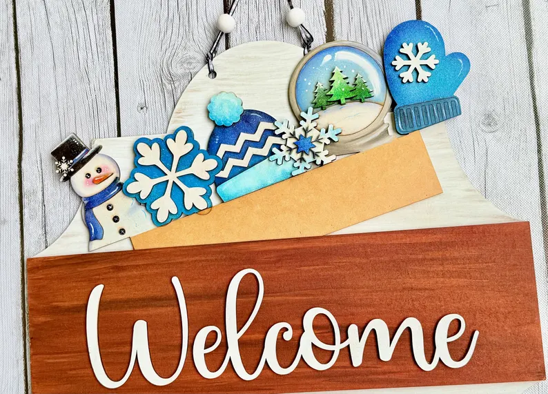 Red Rooster Laser Arts Winter Wonderland Interchangeable Door Hanger Insert features a charming wooden welcome sign adorned with winter-themed ornaments such as a snowman, snowflakes, mittens, and a snowy landscape on a crisp white wooden background. Designed to be fully compatible with the DIY Cute Halloween Interchangeable Door Hanger Insert Kit by Red Rooster Laser Arts, this insert lets you effortlessly update your décor for the season while enjoying year-round versatility.