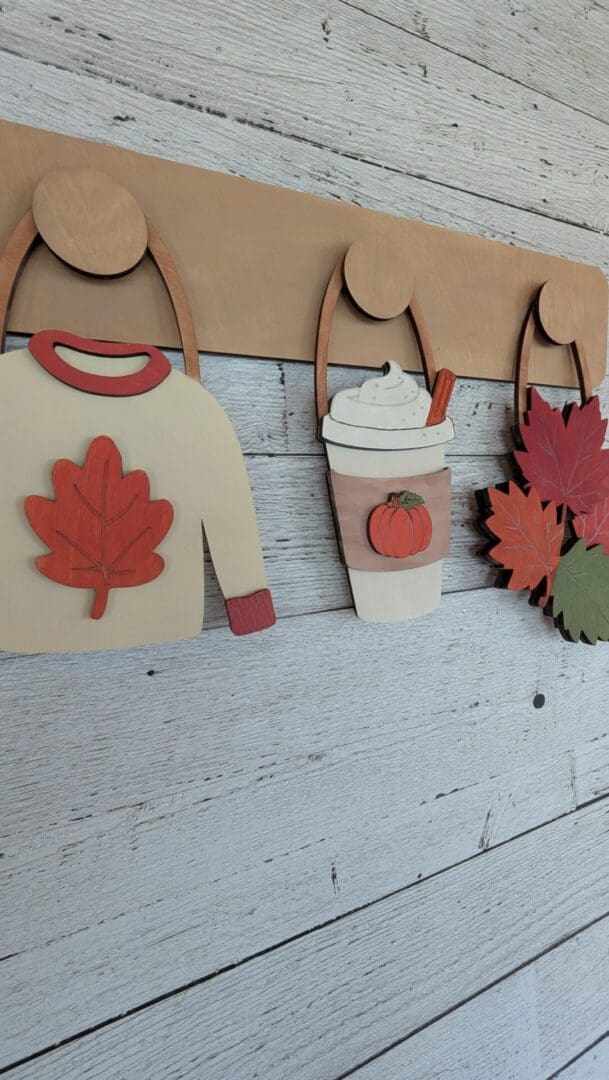 Red Rooster Laser Arts The DIY Interchangeable Wall Peg System Decor - Cozy Fall features a wooden autumn-themed design with a sweater adorned by a red leaf, a pumpkin spice drink complete with straw and cinnamon stick, and three vibrant fall leaves—each hanging on individual hooks. Effortlessly switch out the pieces to refresh your decor for different seasons or create holiday-inspired, patriotic displays.