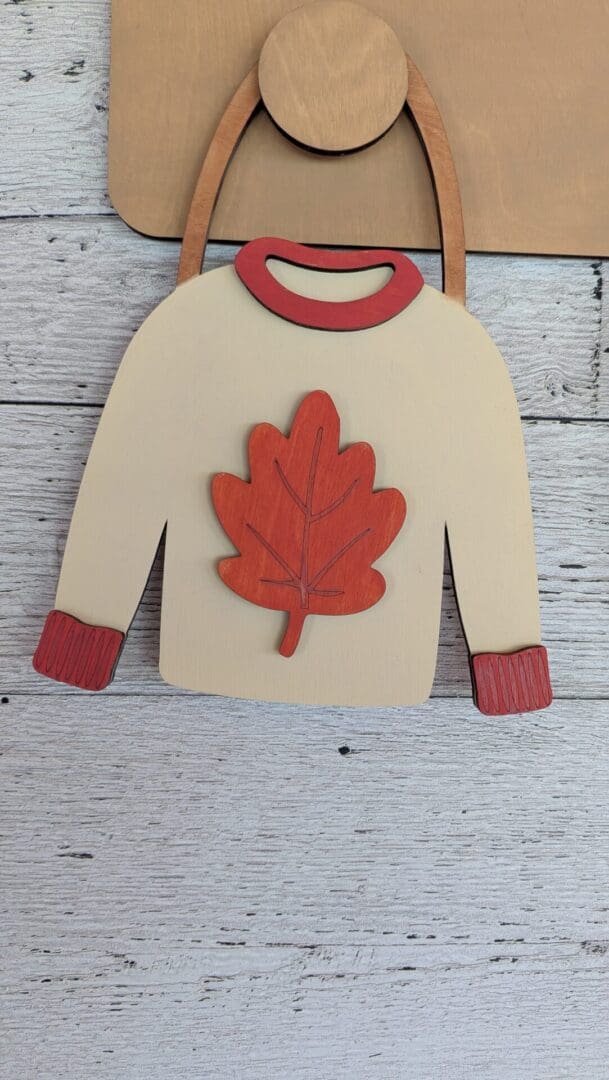 Red Rooster Laser Arts Bring patriotic flair to your DIY Interchangeable Wall Peg System Decor - Cozy Fall with this wooden cutout in the shape of a sweater, detailed with red cuffs and collar and featuring a bold red maple leaf on the chest, displayed against a light wood plank background.