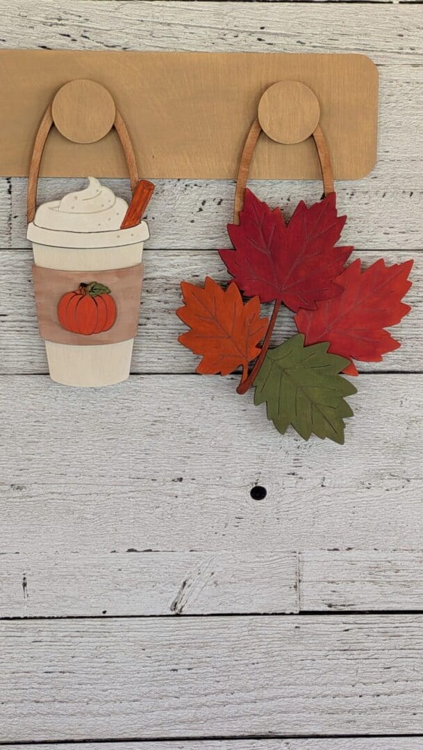 Red Rooster Laser Arts DIY Interchangeable Wall Peg System Decor - Cozy Fall features wooden cutouts of a pumpkin spice latte with whipped cream and a cinnamon stick, along with a bunch of colorful autumn leaves, hanging from hooks on a light wood wall—perfect for easily switching out seasonal or patriotic decor accents.