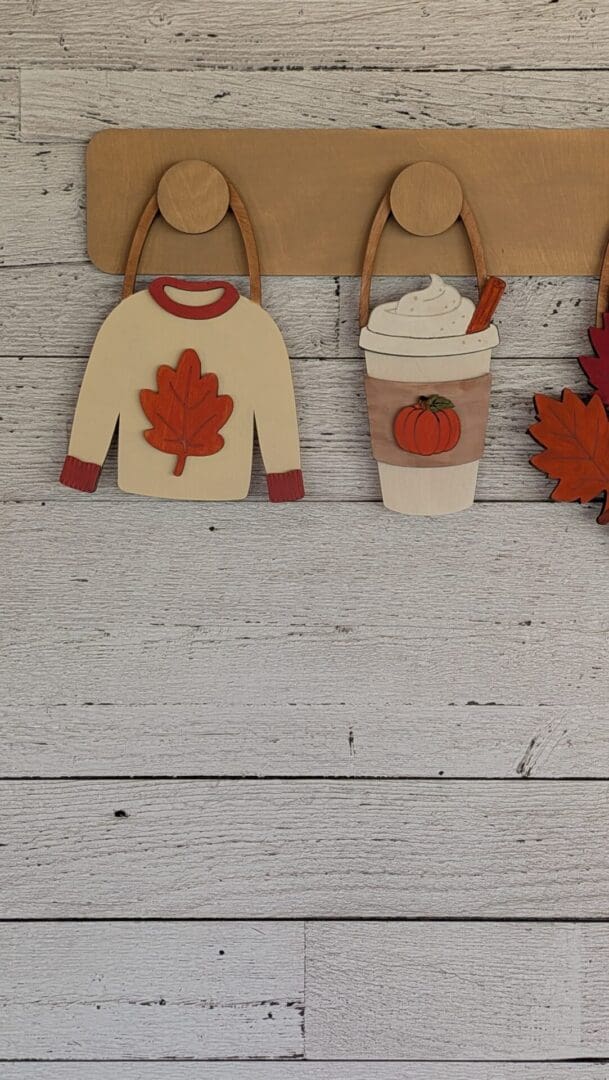 Red Rooster Laser Arts The DIY Interchangeable Wall Peg System Decor - Cozy Fall features wooden cutouts of a sweater with a red leaf and a pumpkin spice drink with whipped cream, hanging on a wooden rack against a white wood wall. This versatile decor is perfect for easily switching out seasonal designs—swap in more patriotic styles as the seasons change!.
