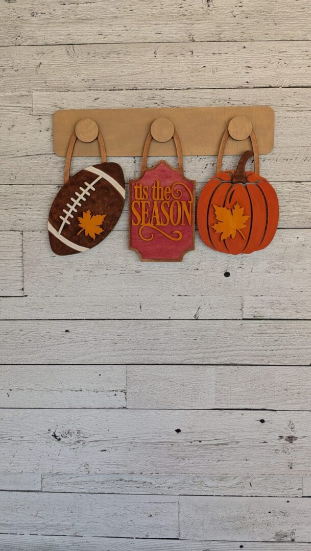 Red Rooster Laser Arts Three wooden fall-themed decorations—a football, a “tis the season” sign, and a pumpkin—hang on hooks against a rustic white wooden wall, each accented with a yellow maple leaf. The DIY Interchangeable Wall Peg System Decor - Fall Football makes it easy to customize your space for fall and showcase your love of football.