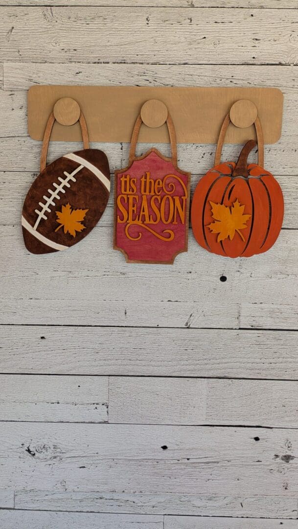 Red Rooster Laser Arts Three wooden fall decorations from the DIY Interchangeable Wall Peg System Decor – Fall Football set hang on hooks: a football with a leaf, a ’tis the season sign, and a pumpkin with a leaf, all displayed against a white wood plank wall.