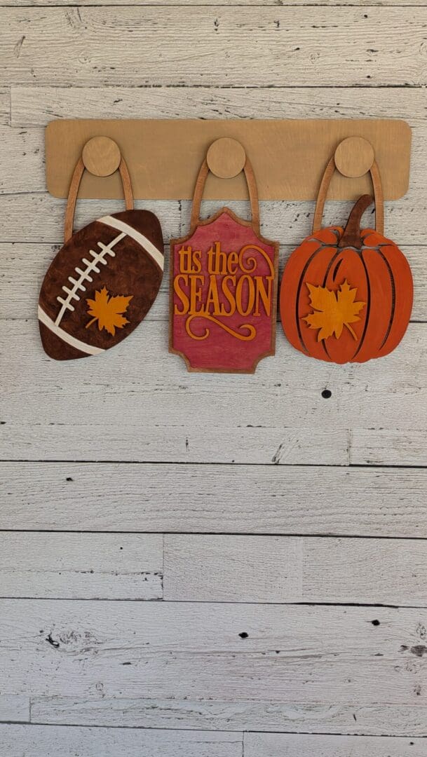 Red Rooster Laser Arts The DIY Interchangeable Wall Peg System Decor - Fall Football features three autumn-inspired decor pieces— a football adorned with a maple leaf, a “tis the season” sign, and a pumpkin with a maple leaf—displayed on wooden hooks against a white wooden wall, making it the perfect way to bring cozy fall football spirit to your home.