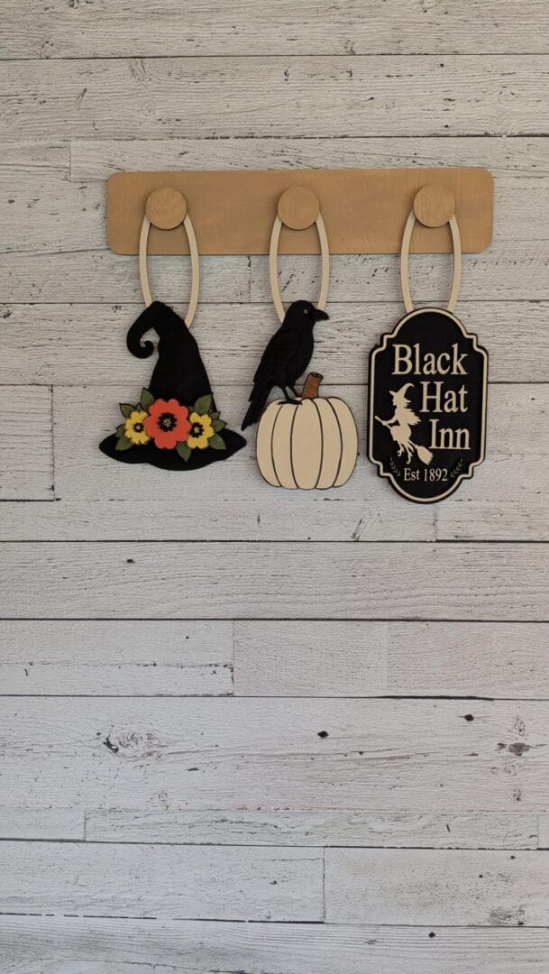 Red Rooster Laser Arts The DIY Interchangeable Wall Peg System Decor - Black Hat Inn Halloween features three wooden hooks displaying a witch hat with flowers, a raven on a white pumpkin, and a Black Hat Inn Est 1892 Halloween sign, creating charming decor you can easily update from Fall to football season—all mounted on a light wooden wall.