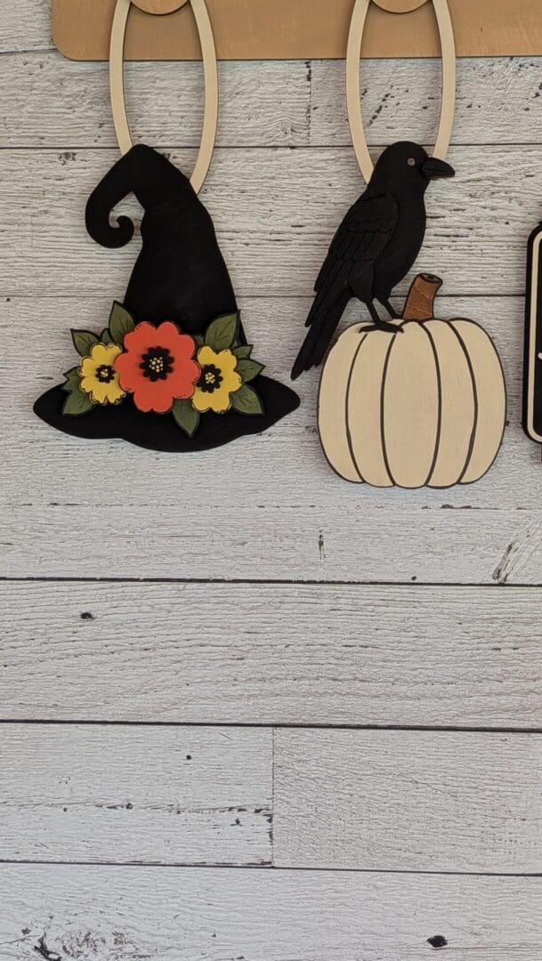 Red Rooster Laser Arts Transform your space for fall with the DIY Interchangeable Wall Peg System Decor - Black Hat Inn Halloween, featuring a black witch hat adorned with flowers and a black crow perched on a white pumpkin—both hanging as stylish wooden decorations on a white wooden wall. Perfect for customizable seasonal décor!.