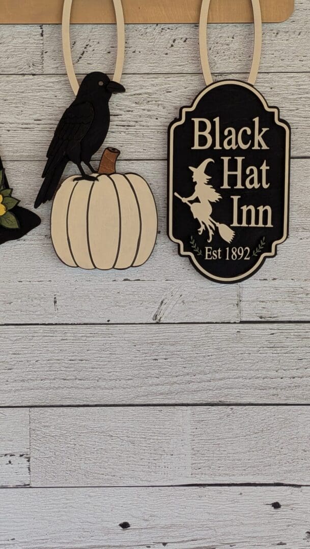 Red Rooster Laser Arts The DIY Interchangeable Wall Peg System Decor – Black Hat Inn Halloween features a wooden sign that reads “Black Hat Inn Est 1892” with a witch silhouette. Beside it, a wooden crow perches atop a white pumpkin, all displayed on a crisp white wooden wall—making it the perfect interchangeable decor piece for transitioning into fall or gearing up for football season.