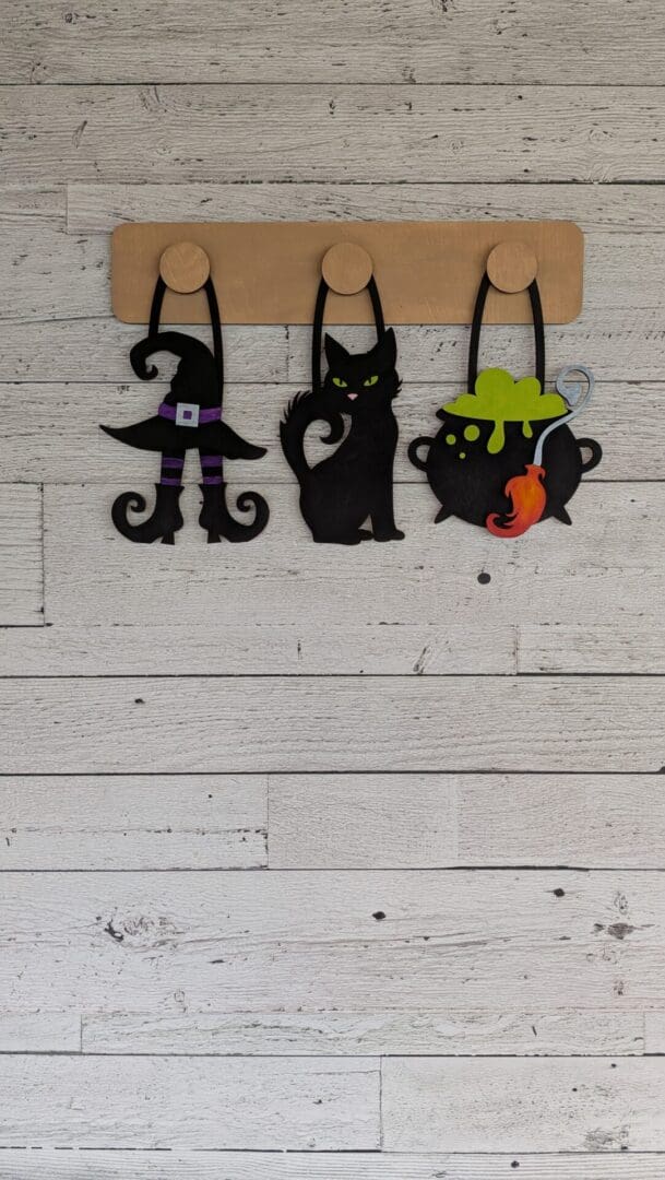 Red Rooster Laser Arts The DIY Interchangeable Wall Peg System Decor - Witchy Halloween features three felt decorations—a witch’s hat and boots, a black cat, and a bubbling cauldron—hanging from wooden hooks on a white wood-paneled wall, creating charming, customizable decor for a witchy Halloween touch.
