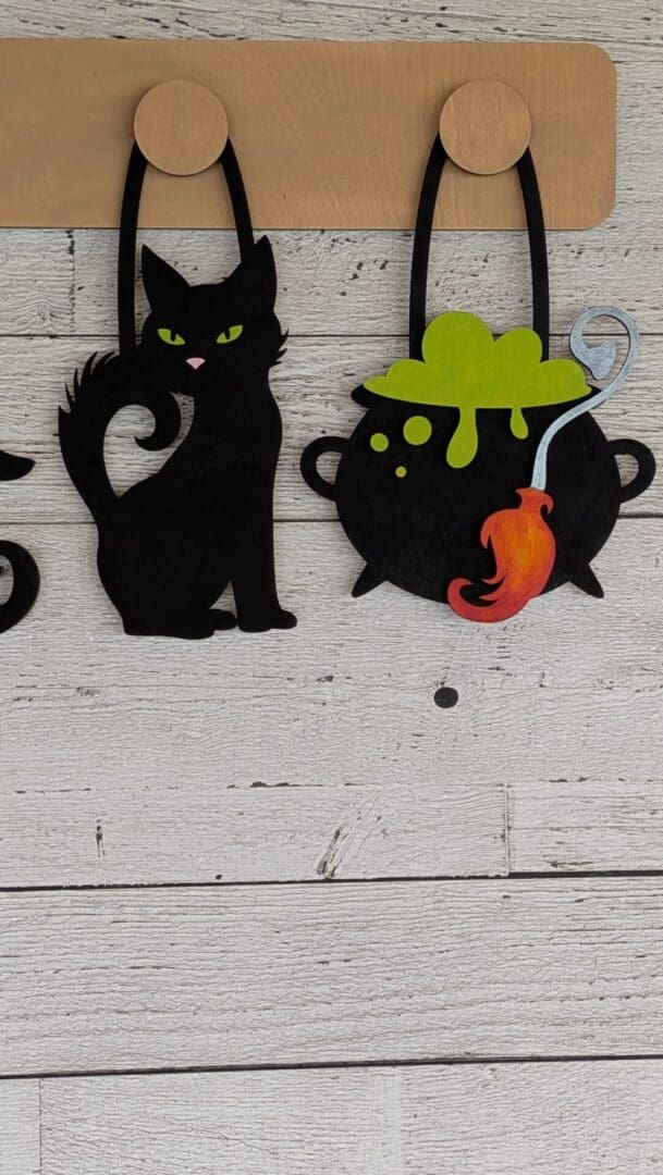 Red Rooster Laser Arts The DIY Interchangeable Wall Peg System Decor - Witchy Halloween features a black cat with green eyes and a bubbling cauldron filled with green liquid and a red ladle, all hanging on a light wood background—perfect for creating witchy Halloween vibes in your space.