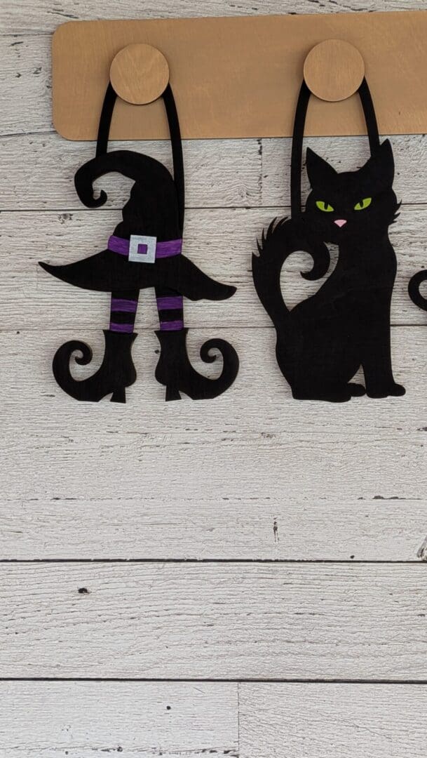 Red Rooster Laser Arts Two DIY Interchangeable Wall Peg System Decor - Witchy Halloween pieces hang on wooden hooks: one features a curled witch hat with boots and a purple belt, while the other showcases a black cat with green eyes. Both are crafted from black material and display whimsical designs.